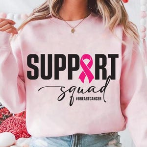 May include: A light pink sweatshirt with the words "SUPPORT" and "squad" in black, and a pink ribbon graphic. The hashtag "#BREASTCANCER" is below the word "squad". The sweatshirt is a crew neck style.