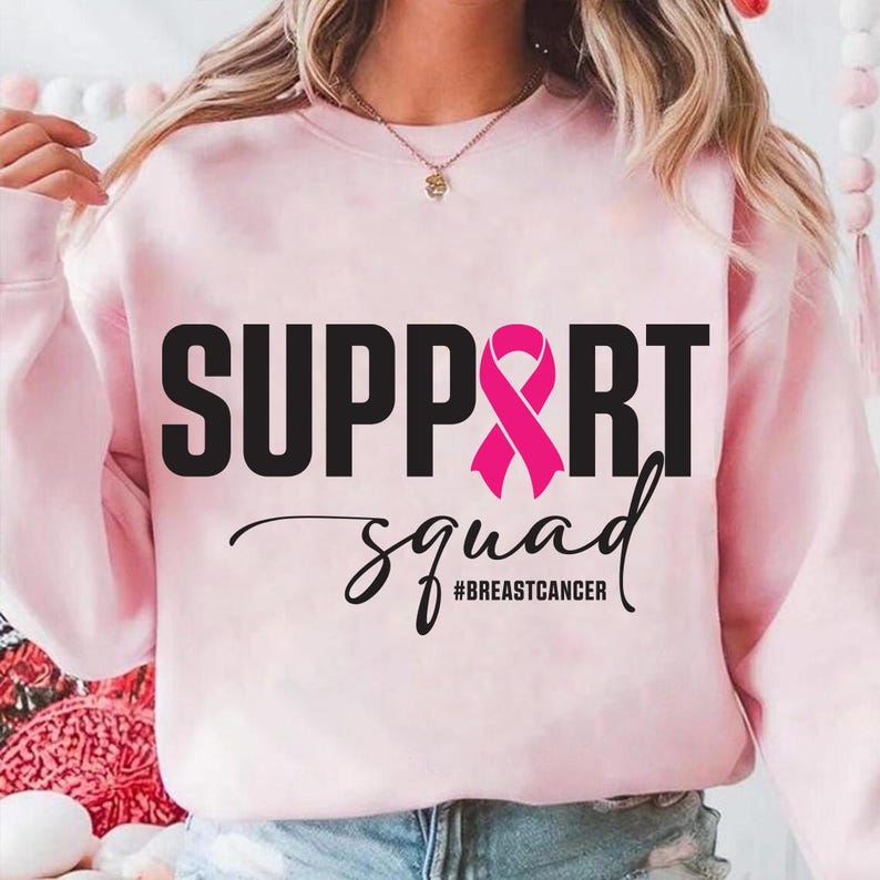 May include: A light pink sweatshirt with the words "SUPPORT" and "squad" in black, and a pink ribbon graphic. The hashtag "#BREASTCANCER" is below the word "squad". The sweatshirt is a crew neck style.