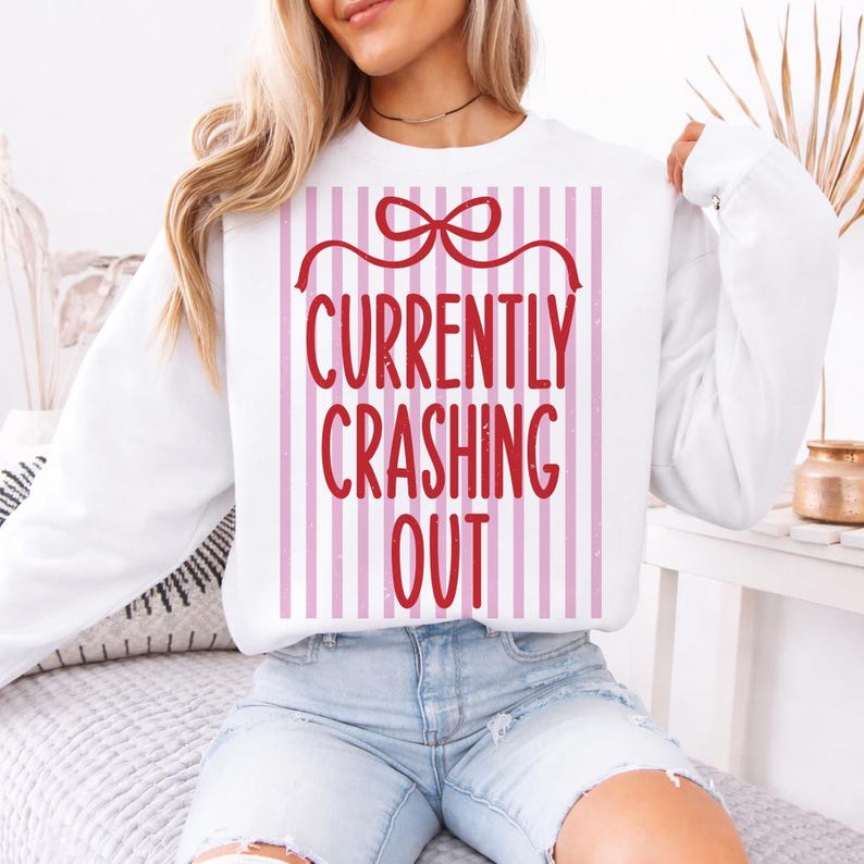 May include: White sweatshirt with the text "CURRENTLY CRASHING OUT" in red, set against a pink and white striped background. The design includes a red bow at the top, creating a gift box appearance.