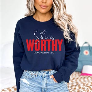 May include: A navy blue sweatshirt with the words "She is WORTHY" in white script and red block letters. The text "PROVERBS 3:1" is below the main text. The sweatshirt is paired with blue jeans.