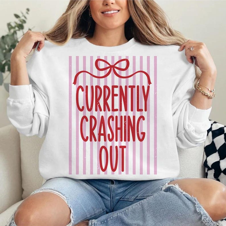 May include: White sweatshirt with long sleeves featuring the text "CURRENTLY CRASHING OUT" in red, set against a pink and white striped background. The design includes a red bow at the top.
