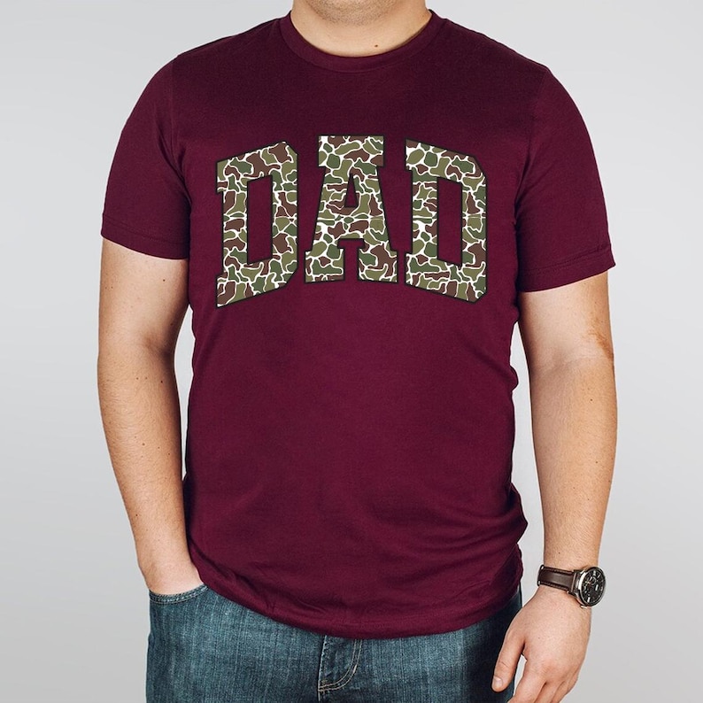 May include: Burgundy t-shirt with a camouflage print "DAD" graphic.