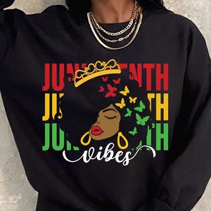 Puede incluir: Sudadera negra con un gráfico que presenta a una mujer negra con una corona, mariposas y las palabras "Juneteenth Vibes" en rojo, amarillo, verde y blanco. Pendientes de aro dorados y un collar de oro también son visibles.