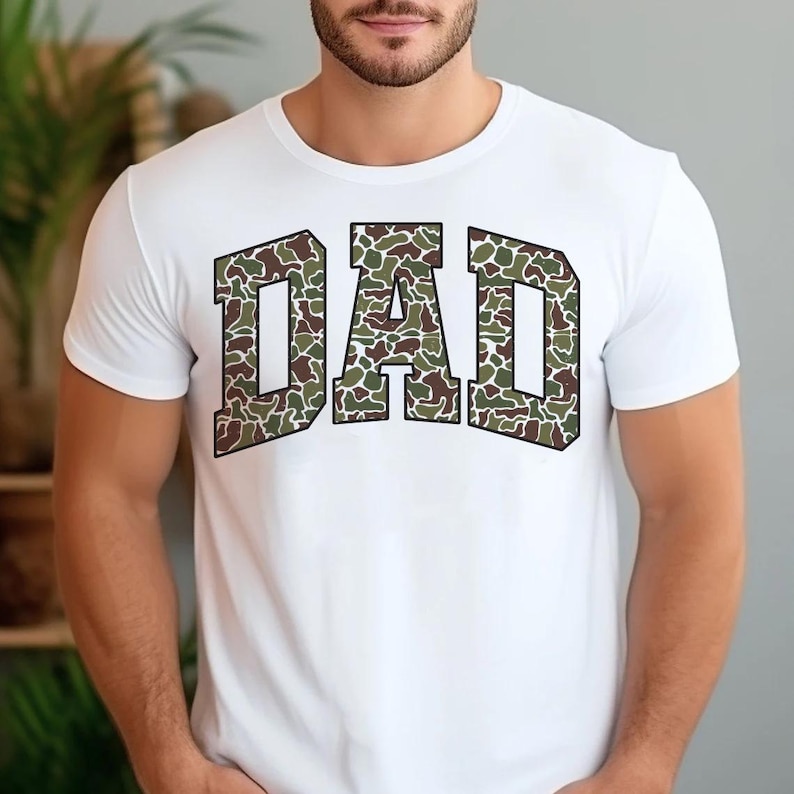 May include: White t-shirt with a camouflage print of the word "DAD" in a bold, blocky font.
