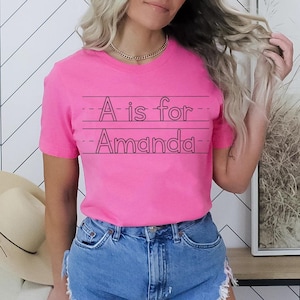 May include: A bright pink t-shirt with the text "A is for Amanda" in black, written on lined paper. The shirt is paired with blue denim shorts. A gold chain necklace is visible.