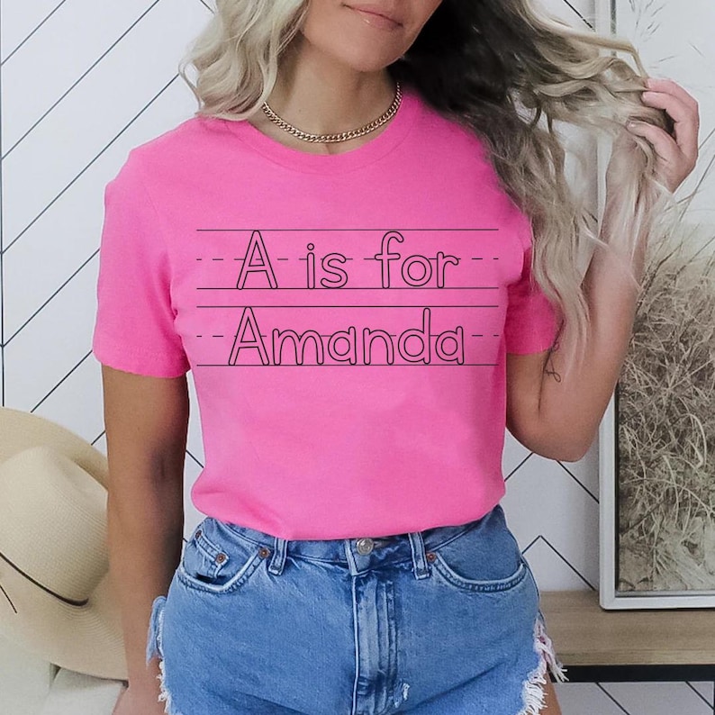 May include: A bright pink t-shirt with the text "A is for Amanda" in black, written on lined paper. The shirt is paired with blue denim shorts. A gold chain necklace is visible.