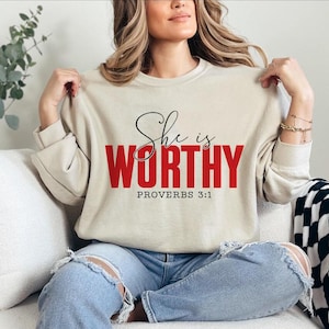 May include: Beige sweatshirt with the text "She is WORTHY" in black script and red block letters, with "PROVERBS 3:1" below. The sweatshirt is long-sleeved and has a crew neck.