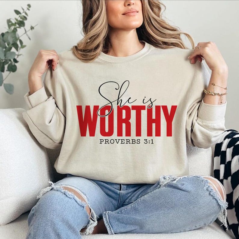 May include: Beige sweatshirt with the text "She is WORTHY" in black script and red block letters, with "PROVERBS 3:1" below. The sweatshirt is long-sleeved and has a crew neck.