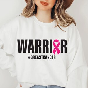 May include: White sweatshirt with the word "WARRIOR" in bold black letters and a pink ribbon graphic. Below the word "WARRIOR" is the hashtag "#BREASTCANCER". The sweatshirt is a crew neck style.