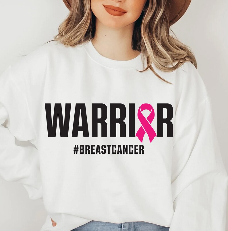 May include: White sweatshirt with the word "WARRIOR" in bold black letters and a pink ribbon graphic. Below the word "WARRIOR" is the hashtag "#BREASTCANCER". The sweatshirt is a crew neck style.