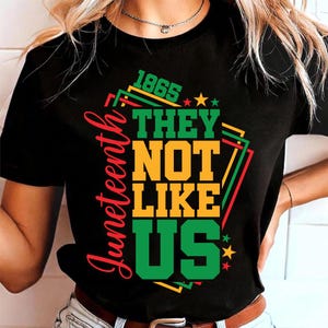May include: Black t-shirt with the text "Juneteenth" in red script on the left side. The shirt features the words "THEY NOT LIKE US" in yellow and green, with the year "1865" above. Stars and geometric shapes in red, green, and yellow complete the design.