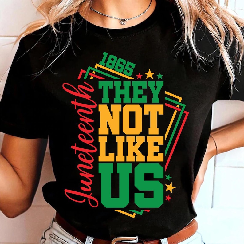 May include: Black t-shirt with the text "Juneteenth" in red script on the left side. The shirt features the words "THEY NOT LIKE US" in yellow and green, with the year "1865" above. Stars and geometric shapes in red, green, and yellow complete the design.