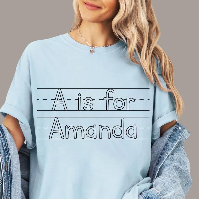 May include: Light blue t-shirt with the text "A is for Amanda" in black. The text is written in a school-style font, with lines above and below the text. The shirt has short sleeves.