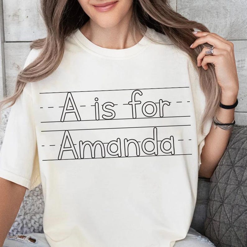 May include: Cream-colored t-shirt with the text "A is for Amanda" in black, written in a school notebook style. The shirt has a crew neck and short sleeves.