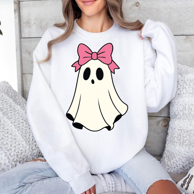 K&ouml;nnte beinhalten: Wei&szlig;es Sweatshirt mit einem Cartoon-Geist-Design. Der Geist hat eine rosa Schleife, schwarze Augen und schwarze Details am Saum. Ein l&auml;ssiges, bequemes Oberteil, passend f&uuml;r Halloween oder den t&auml;glichen Gebrauch.