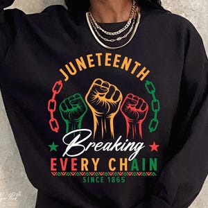 Könnte beinhalten: Schwarzes Sweatshirt mit einem Grafikdesign zur Feier des Juneteenth. Das Design zeigt drei erhobene Fäuste in Grün, Orange und Rot, umgeben von einer Kette. Der Text lautet "Juneteenth", "Breaking Every Chain" und "Since 1865."