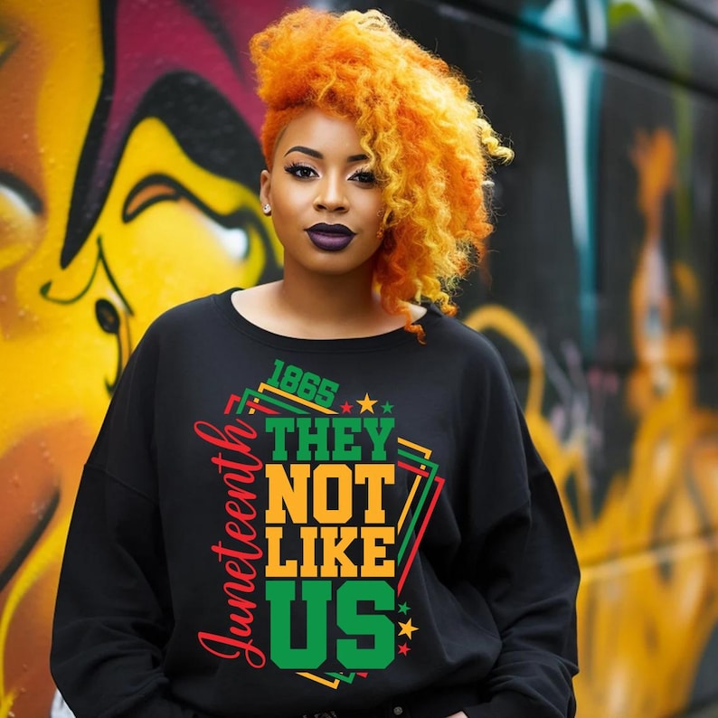 May include: Black sweatshirt with the text "Juneteenth 1865 THEY NOT LIKE US" in green, orange, and red. The sweatshirt is worn by a person with orange and yellow hair and dark lipstick. The background features colorful graffiti.