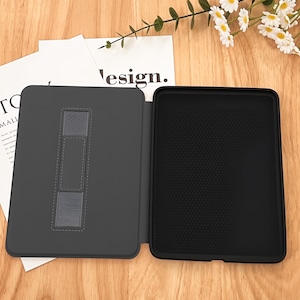 May include: A dark gray tablet case, open to display the interior. The left side features a stand, while the right side has a textured black surface. The word "design" is partially visible on a white paper in the background. The case is on a wooden surface.