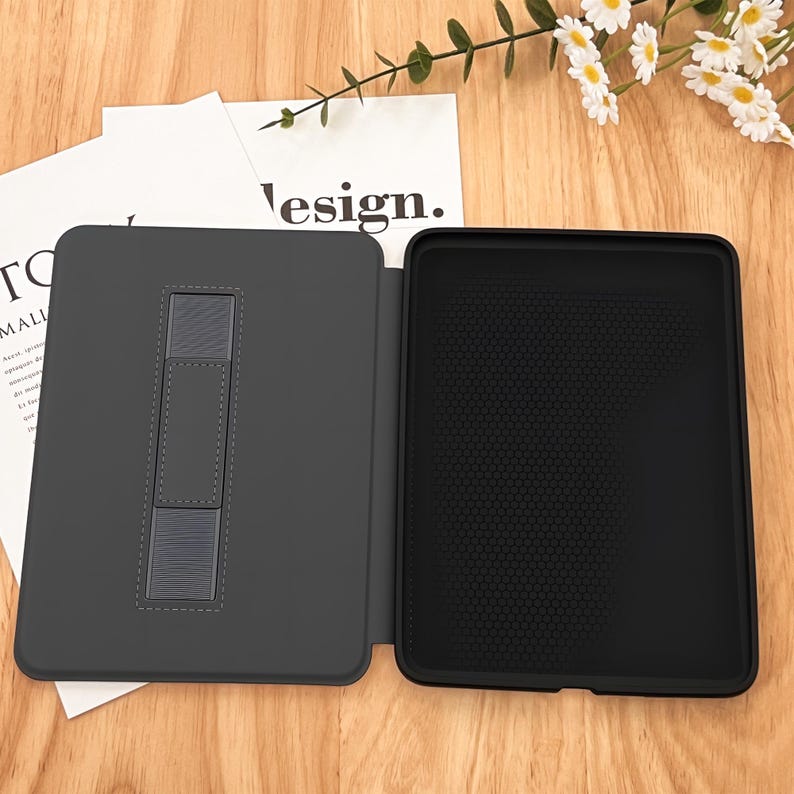 May include: A dark gray tablet case, open to display the interior. The left side features a stand, while the right side has a textured black surface. The word "design" is partially visible on a white paper in the background. The case is on a wooden surface.