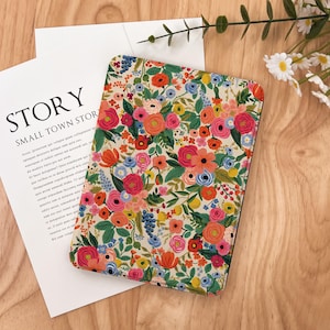 May include: A floral patterned notebook cover with a cream background and a variety of colorful flowers. The cover is open, and the text "STORY SMALL TOWN STORY" is visible on a white paper. The notebook is on a wooden surface.