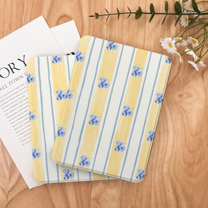 May include: Two rectangular cases with a yellow and white striped pattern and blue blueberry illustrations. The cases are on a wooden surface with a white paper and a sprig of flowers.