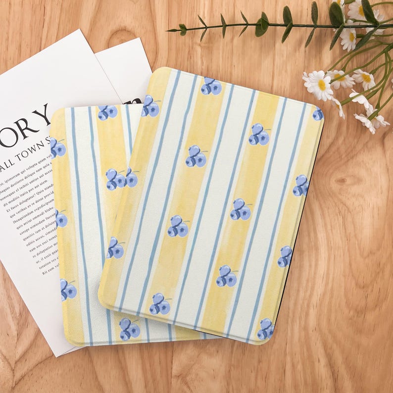 May include: Two rectangular cases with a yellow and white striped pattern and blue blueberry illustrations. The cases are on a wooden surface with a white paper and a sprig of flowers.