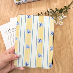May include: A patterned e-reader cover featuring a yellow and white striped background with blue blueberry illustrations. The cover is held over a wooden surface, with a book and flowers in the background.