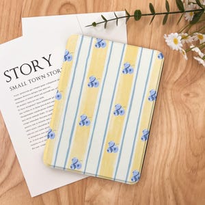 Puede incluir: Una funda para tableta con un diseño de rayas amarillas y blancas, adornada con ilustraciones de arándanos azules. La funda está sobre una superficie de madera, con un papel blanco y el texto "STORY SMALL TOWN STOR".