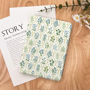 May include: A tablet case featuring a floral pattern with a white backdrop and a repeating design of blue, green, and yellow flowers. The case is on a wooden surface next to a white paper with the word "STORY" printed on it.