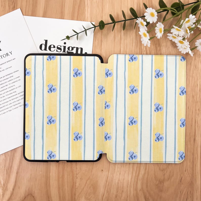 May include: An open tablet case with a yellow and white striped pattern, adorned with small blue floral designs. The case is black-edged and lies on a wooden surface, with a sprig of white flowers and the word "design" visible.