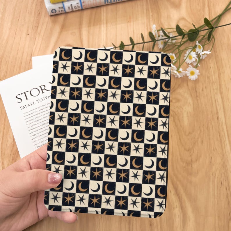 May include: A tablet case with a celestial design of moons and stars in a checkerboard pattern. The case has a black, cream, and gold colour scheme. The case is held by a person's hand. The word "STOR" is visible on a nearby book.