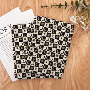 May include: Two notebooks with a celestial pattern of gold stars and crescent moons on a black and cream checkered background. The notebooks are on a wooden surface with a white paper and flowers.