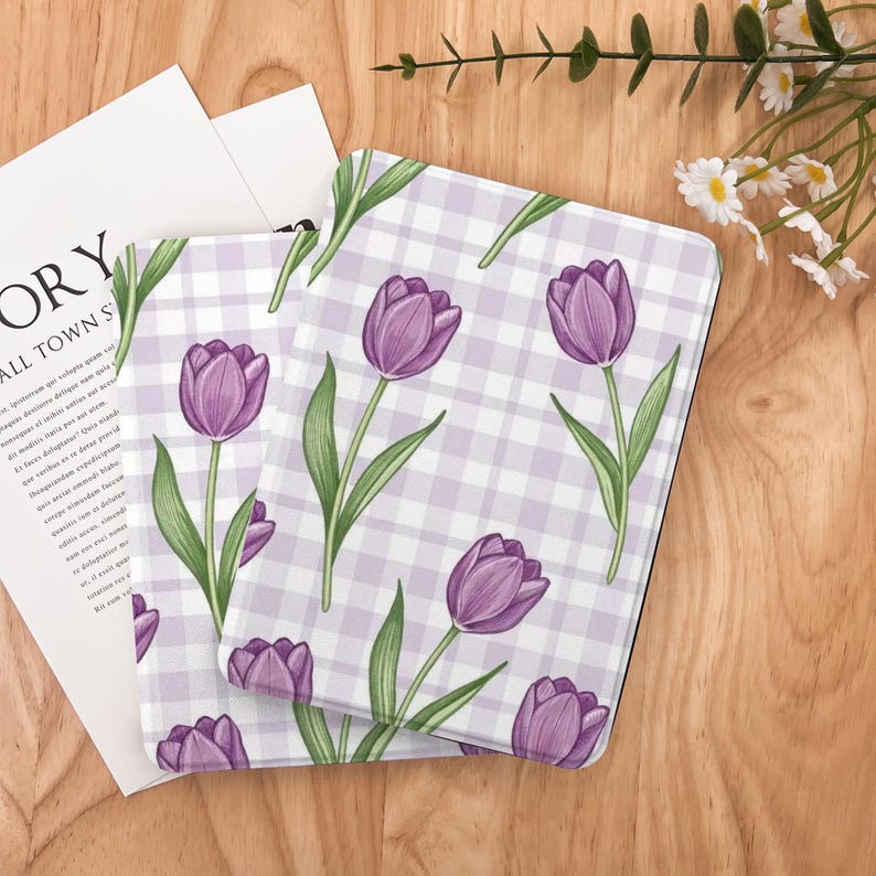 May include: Two square coasters with a purple tulip floral pattern on a light purple and white checkered background. The coasters are placed on a wooden surface, with a white paper and some flowers in the background.