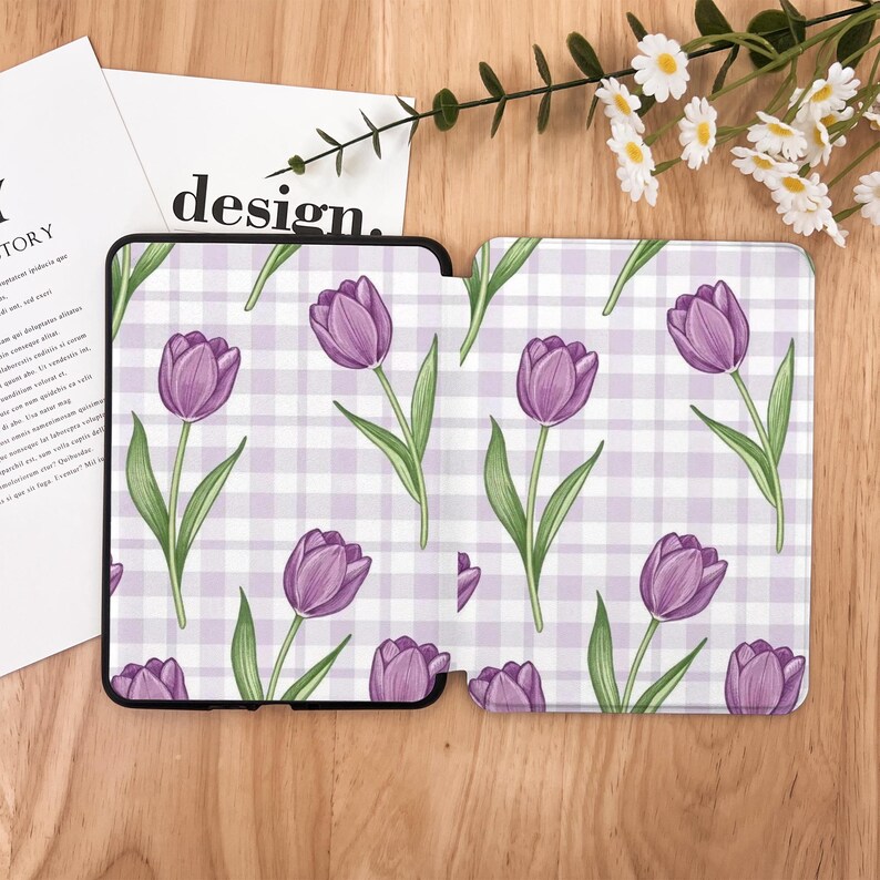 May include: A phone case and a tablet case with a purple tulip pattern on a white and light purple gingham background. The cases are black and the design includes green stems and leaves. The word "design" is visible on a white paper.