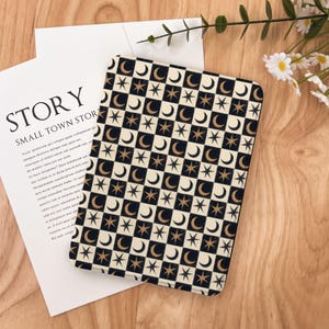 Pode incluir: Uma capa retangular com um padrão celestial de luas e estrelas pretas e douradas sobre um fundo xadrez creme. A capa está sobre uma superfície de madeira com um papel branco com a palavra "STORY" impressa.