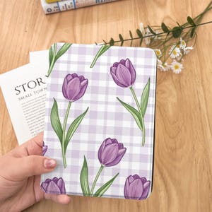 May include: A tablet case featuring a purple tulip pattern on a white and lavender gingham background. The floral design includes green stems and leaves. The case is held in a person's hand, with a book and flowers in the background.
