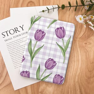 May include: A rectangular case featuring a purple tulip floral pattern on a light purple and white checkered background. The case is on a wooden surface with a white paper and the text "STORY SMALL TOWN STOR".