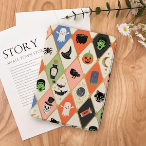 May include: A Halloween-themed tablet case with a repeating diamond pattern. The design includes ghosts, pumpkins, bats, and a black cat. The case is on a wooden surface. The word "TREAT" is visible.