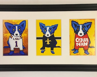 May include: Three framed prints of a blue dog wearing different outfits. The dog on the left is wearing a white and purple LSU jersey with the number 1. The dog in the middle is wearing a black and gold Saints jersey with a fleur-de-lis. The dog on the right is wearing a red shirt with the words "Cajun Man".