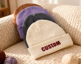 May include: A stack of four ribbed knit beanies in cream, charcoal gray, lavender, and brown. The cream-colored beanie features the word CUSTOM embroidered in dark red capital letters on the front cuff of the hat.