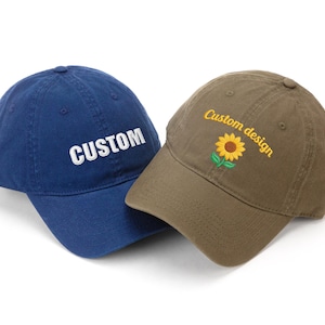 May include: Two baseball caps. One is navy blue with the word "CUSTOM" embroidered in white. The other is olive green with a sunflower and the words "Custom design" embroidered in yellow.