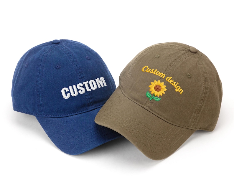 May include: Two baseball caps. One is navy blue with the word "CUSTOM" embroidered in white. The other is olive green with a sunflower and the words "Custom design" embroidered in yellow.