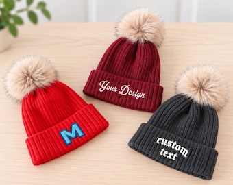 May include: Three ribbed knit beanies with faux fur pom-poms. One is bright red with a blue and light blue letter M patch. One is dark red with white embroidered text reading Your Design. One is dark gray with white embroidered text custom text.