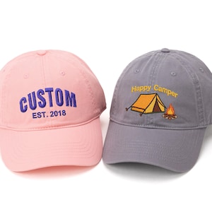 May include: Two baseball caps. One is pink with "CUSTOM EST. 2018" embroidered in blue. The other is gray with "Happy Camper" and a tent and campfire embroidered in yellow and orange. Both have curved brims.