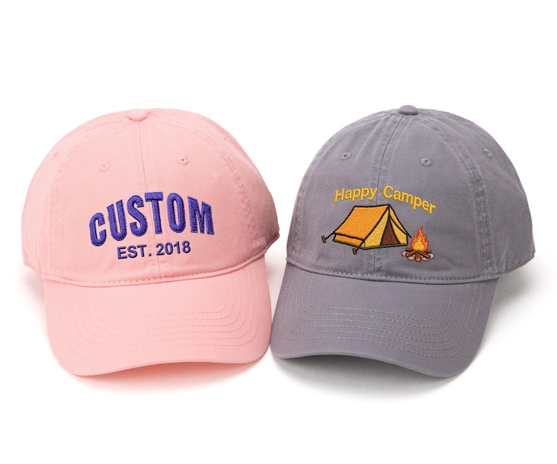 May include: Two baseball caps. One is pink with "CUSTOM EST. 2018" embroidered in blue. The other is gray with "Happy Camper" and a tent and campfire embroidered in yellow and orange. Both have curved brims.