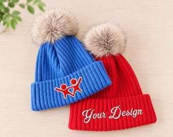 May include: Two ribbed knit beanies with faux fur pom-poms. The blue beanie features an embroidered red stick figure graphic. The red beanie features the white embroidered text Your Design on the folded cuff.