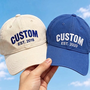 Könnte beinhalten: Eine beige Baseballkappe mit blauem Sticktext CUSTOM EST. 2018 und eine blaue Baseballkappe mit weißem Sticktext CUSTOM EST. 2020. Beide Kappen verfügen über eine Sechs-Panel-Konstruktion mit einem gebogenen Schirm und einem Knopf oben.