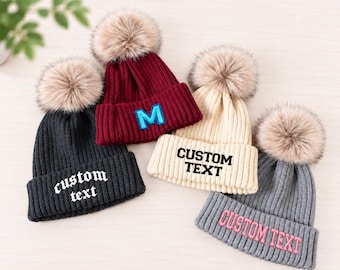 May include: Four ribbed knit beanies with faux fur pom-poms. The black beanie features white text reading custom text. The maroon beanie has a blue letter M. The cream beanie displays black text reading CUSTOM TEXT. The gray beanie has pink text reading CUSTOM TEXT.