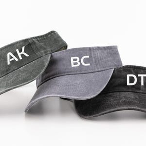 May include: Three sun visors in charcoal, slate grey, and black with a distressed, washed-out texture. Each visor features white capital letters printed on the front panel: AK on the charcoal visor, BC on the slate grey visor, and DT on the black visor.