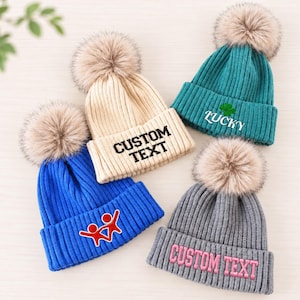 May include: Four ribbed knit beanies with faux fur pom-poms. One beige beanie reads CUSTOM TEXT in black. One teal beanie features a green clover and the word LUCKY. One blue beanie has a red graphic. One gray beanie reads CUSTOM TEXT in pink.